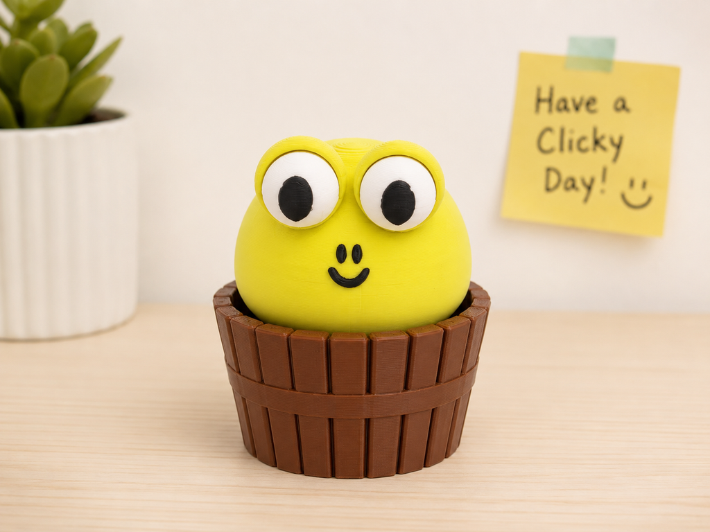 Cute green monster mechanical clicker desk fidget toy. A green monster clicker item with large cartoon eyes and a black smiley face, sitting inside a brown basket-like cup on a light wood surface. A small potted plant is blurred on the left, and a yellow sticky note reading 'Have a Clicky Day! :)' is on the right.