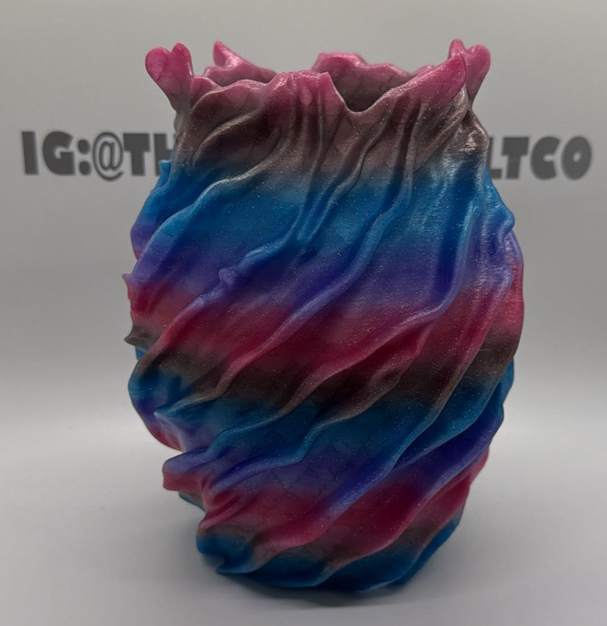 A sculptural, organic-shaped vase with a wavy, ruffled rim, featuring a marbled pattern of blue, pink, purple, and dark gray. The vase is photographed on a white studio background with a faint watermark logo in the background.