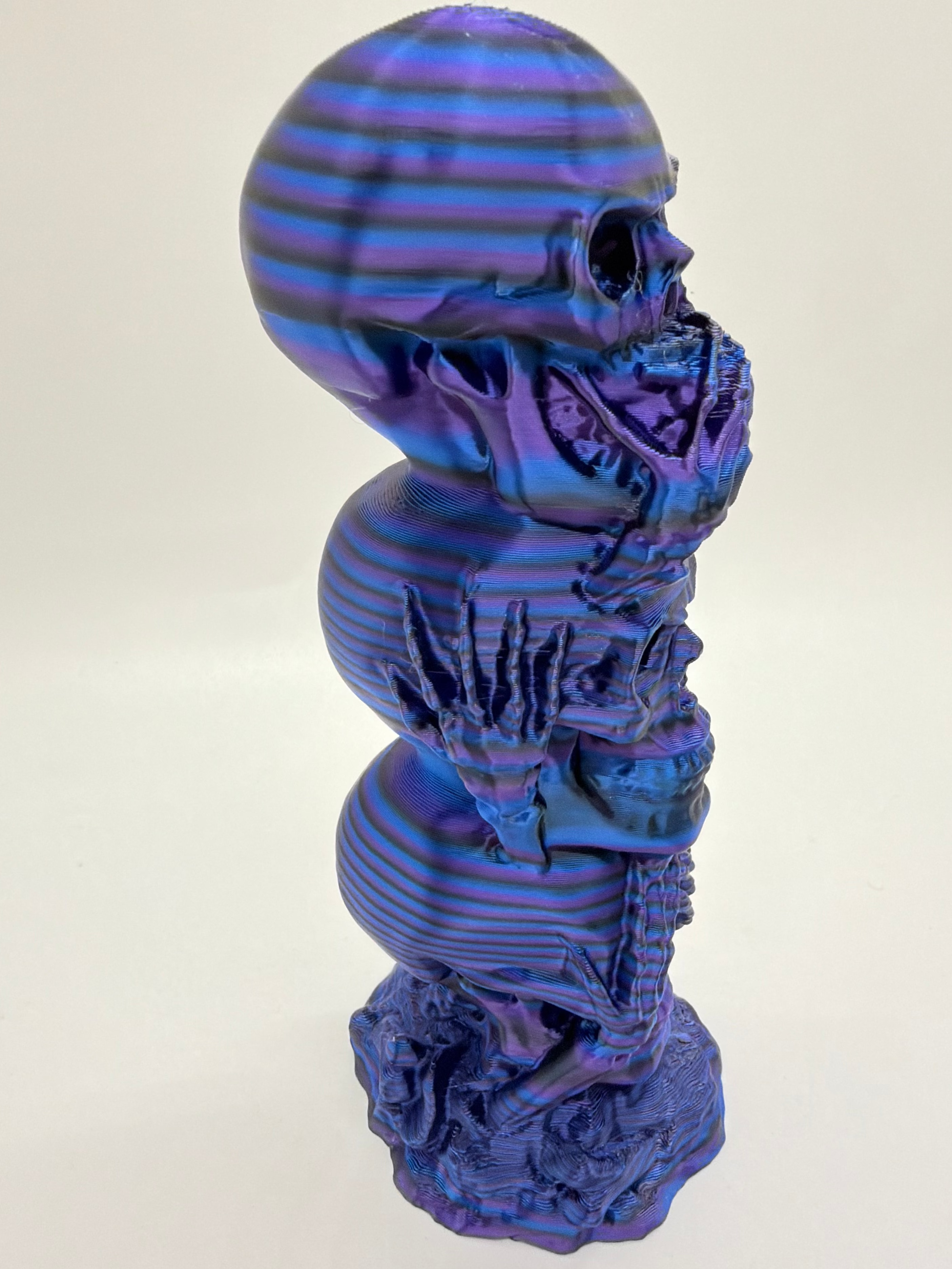 A sculptural figurine of stacked skulls with a glossy, iridescent finish that shifts between purple, blue, and violet. The object is photographed against a plain, light off-white background.