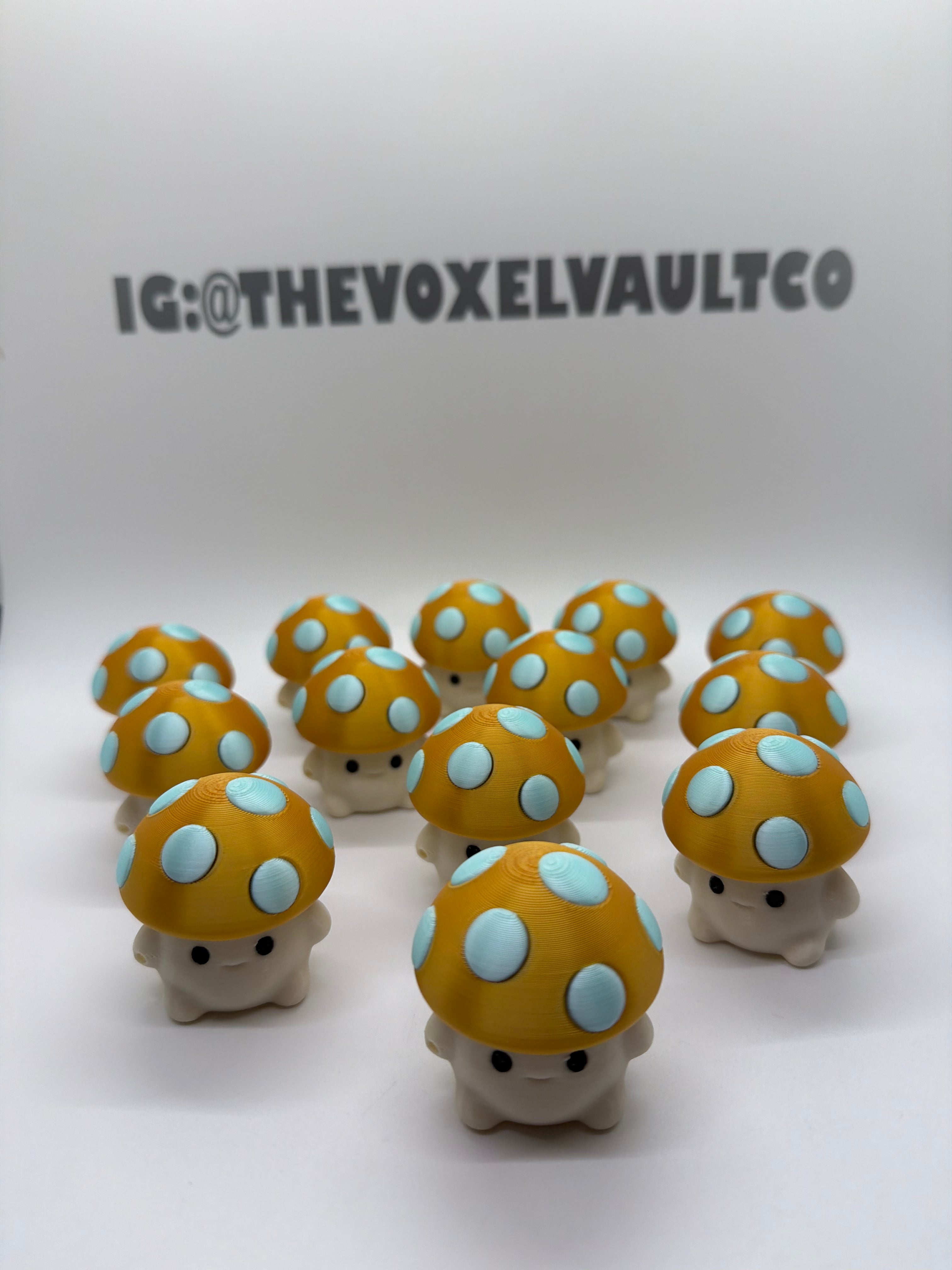 A group of small mushroom-shaped figurines with yellow-orange caps, light blue spots, and cream-colored feet, arranged on a white surface against a white backdrop. The text 'IG:@THEVOXELVAULTCO' is visible on the wall behind them.